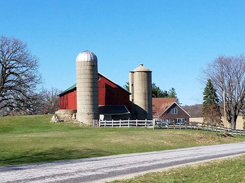 Rural countryside picture of a barn with 2 silos and the road in front.